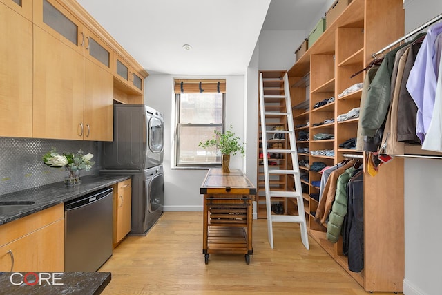 kitchen featuring light wood finished floors, decorative backsplash, stainless steel dishwasher, stacked washing maching and dryer, and dark stone counters