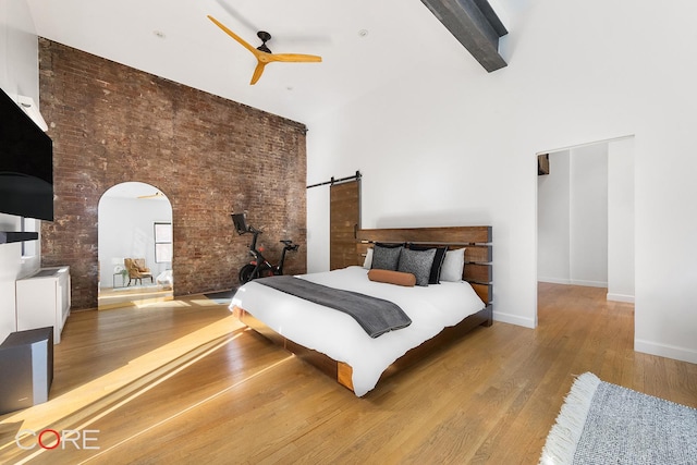 bedroom with a barn door, arched walkways, brick wall, wood finished floors, and a high ceiling