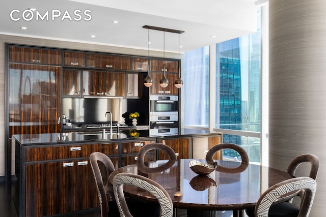 kitchen with dark brown cabinets, modern cabinets, stainless steel double oven, dark countertops, and a kitchen island with sink