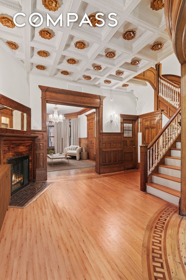 unfurnished living room with a warm lit fireplace, coffered ceiling, stairway, wainscoting, and light wood-type flooring