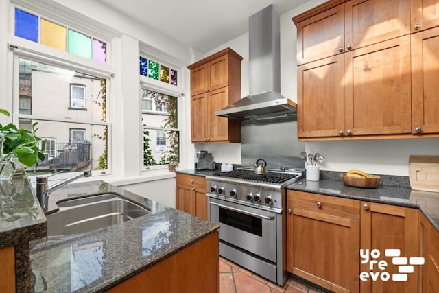 kitchen featuring dark stone counters, wall chimney exhaust hood, brown cabinets, stainless steel stove, and a sink