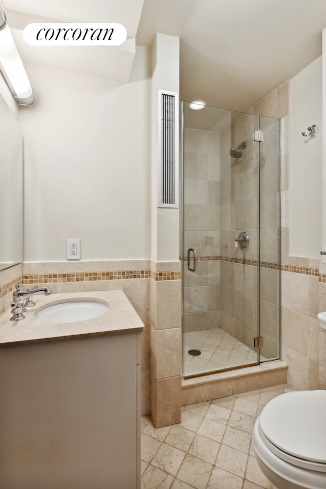 bathroom with tile walls, a shower stall, toilet, wainscoting, and vanity