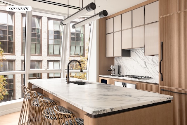 kitchen with light stone counters, a center island with sink, stainless steel gas stovetop, backsplash, and a sink