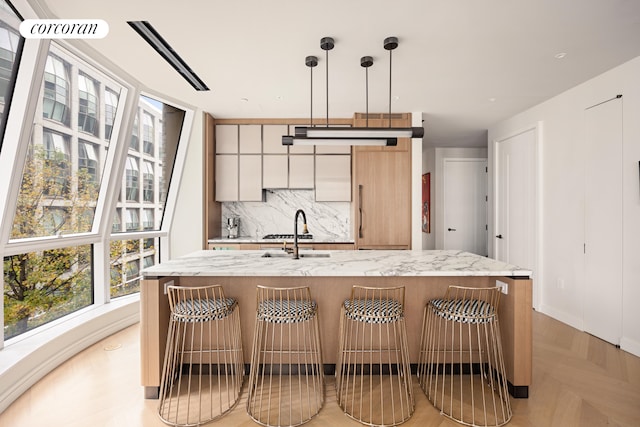 kitchen featuring a center island with sink, a sink, tasteful backsplash, light stone countertops, and hanging light fixtures