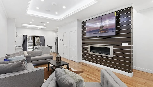 living area featuring a tray ceiling, crown molding, recessed lighting, light wood-style floors, and a glass covered fireplace