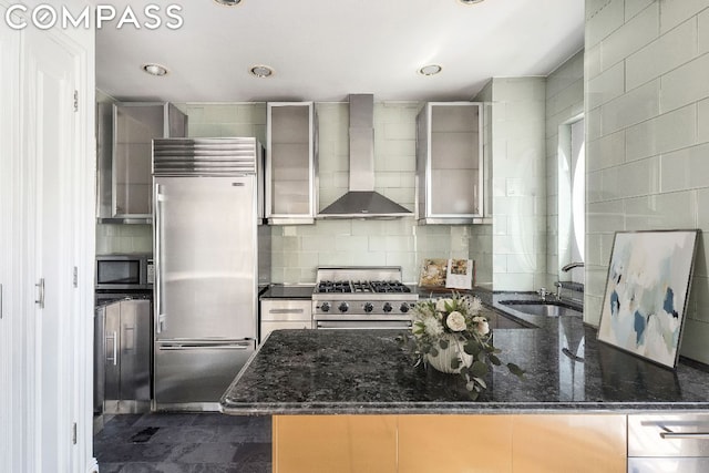 kitchen with backsplash, dark stone counters, high end appliances, wall chimney exhaust hood, and sink