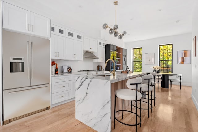 kitchen with wall chimney exhaust hood, white cabinets, stainless steel built in refrigerator, and an island with sink