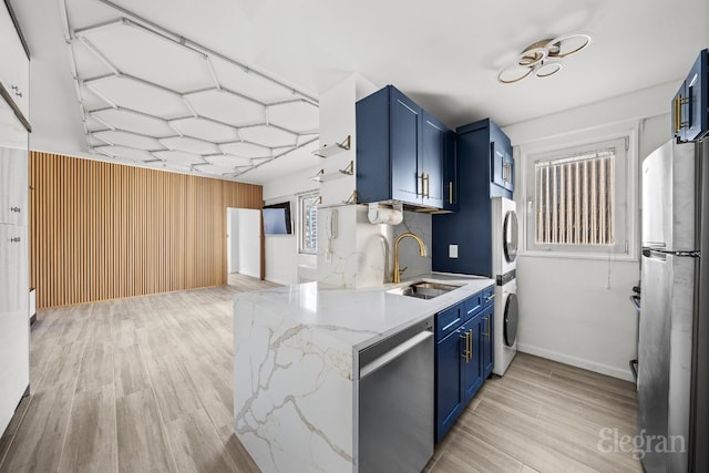 kitchen with sink, blue cabinetry, stainless steel appliances, stacked washer / drying machine, and light stone countertops