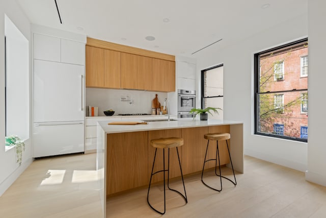 kitchen featuring sink, paneled built in refrigerator, backsplash, an island with sink, and white cabinets