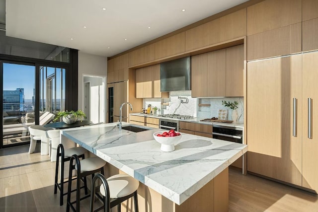 kitchen featuring sink, backsplash, stainless steel appliances, a center island with sink, and wall chimney exhaust hood