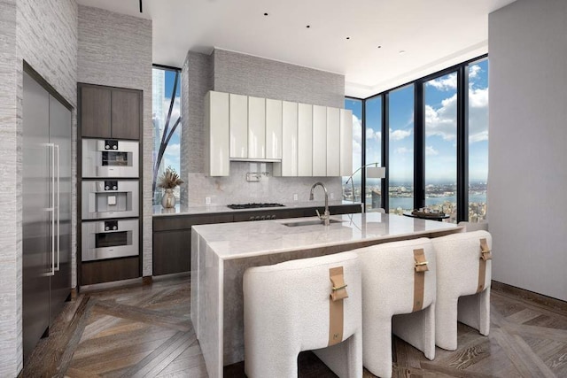 kitchen with white cabinetry, sink, a wall of windows, dark parquet flooring, and a center island with sink