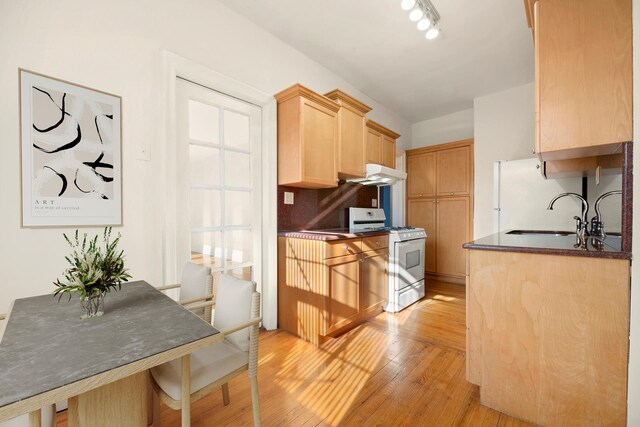 kitchen with light brown cabinetry, under cabinet range hood, a sink, dark countertops, and white gas range oven