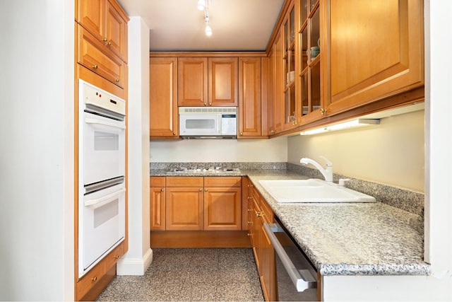 kitchen with light stone counters, white appliances, a sink, brown cabinetry, and glass insert cabinets