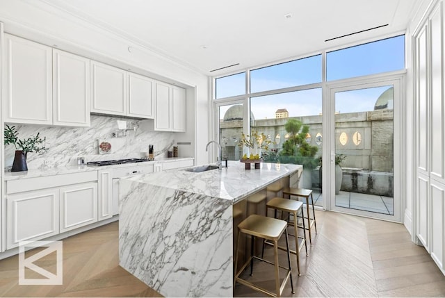 kitchen featuring sink, white cabinetry, backsplash, light stone countertops, and a center island with sink