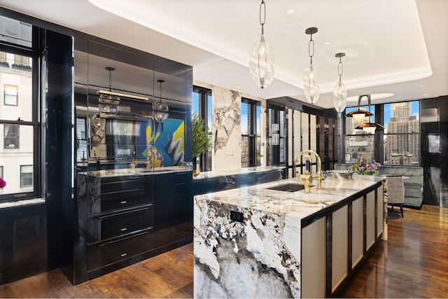 kitchen featuring a tray ceiling, floor to ceiling windows, dark wood-type flooring, a sink, and an island with sink