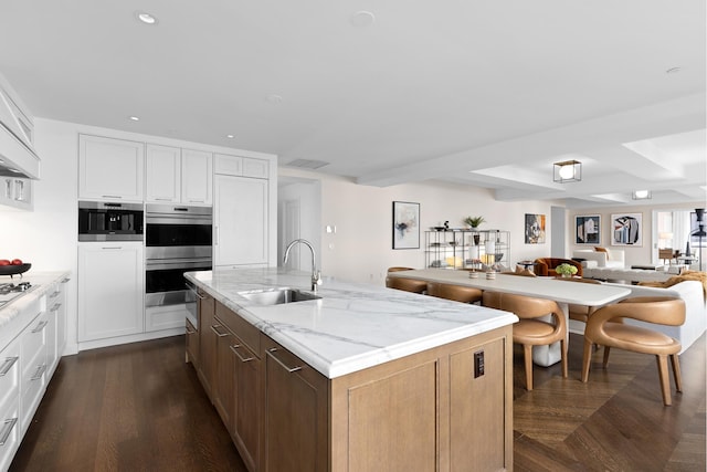 kitchen featuring stainless steel double oven, a sink, open floor plan, a center island with sink, and dark wood-style flooring