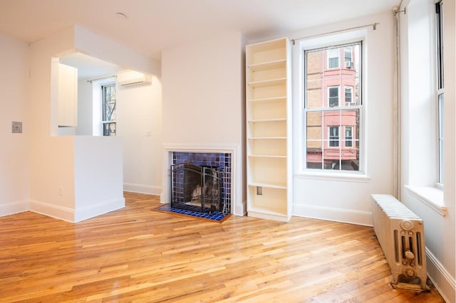 unfurnished living room with a wall unit AC, a tiled fireplace, light wood-type flooring, and radiator heating unit