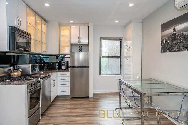 kitchen with a wall unit AC, wood finished floors, dark stone counters, appliances with stainless steel finishes, and white cabinetry