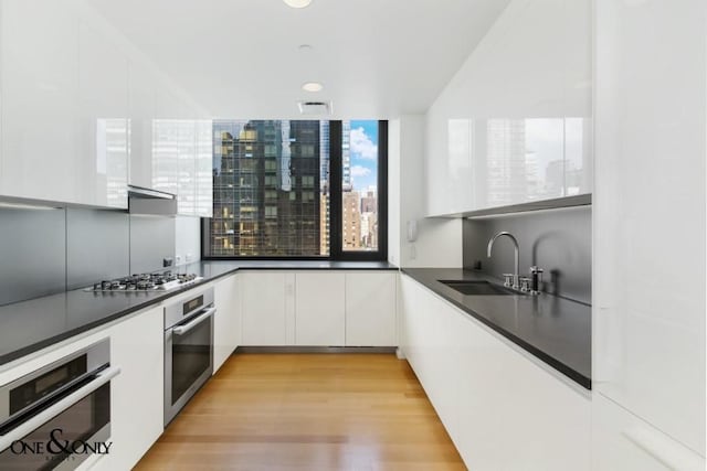 kitchen with appliances with stainless steel finishes, ventilation hood, white cabinetry, sink, and light hardwood / wood-style flooring