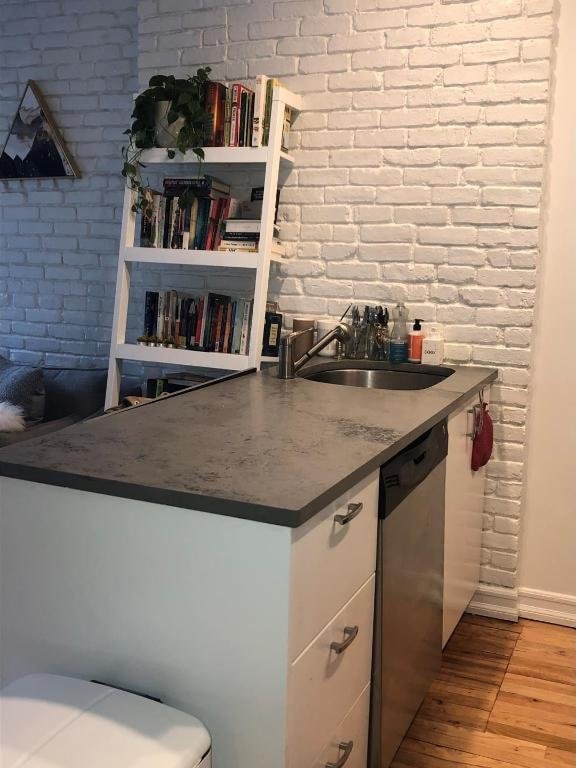 kitchen with a sink, brick wall, wood finished floors, and stainless steel dishwasher