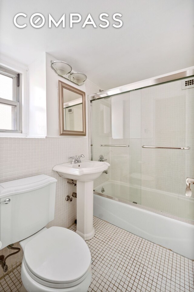 bathroom featuring tile patterned flooring, toilet, wainscoting, combined bath / shower with glass door, and tile walls