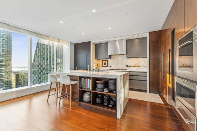 kitchen with light hardwood / wood-style flooring, wall chimney range hood, tasteful backsplash, a breakfast bar, and a center island with sink