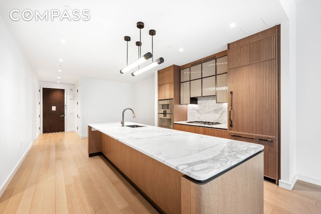 kitchen with sink, light wood-type flooring, an island with sink, pendant lighting, and decorative backsplash