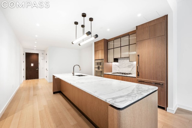 kitchen featuring an island with sink, sink, decorative backsplash, hanging light fixtures, and light wood-type flooring