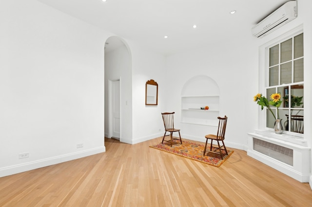 sitting room with baseboards, a wall mounted AC, built in shelves, arched walkways, and light wood finished floors