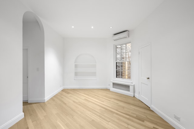 unfurnished room featuring a wall unit AC, arched walkways, built in shelves, light wood finished floors, and recessed lighting