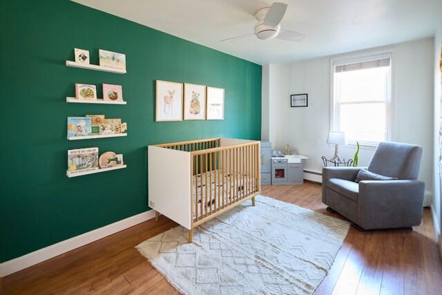 bedroom with ceiling fan, a baseboard radiator, hardwood / wood-style floors, and a nursery area