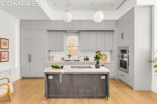 kitchen with a center island with sink, gray cabinets, backsplash, hanging light fixtures, and light hardwood / wood-style flooring