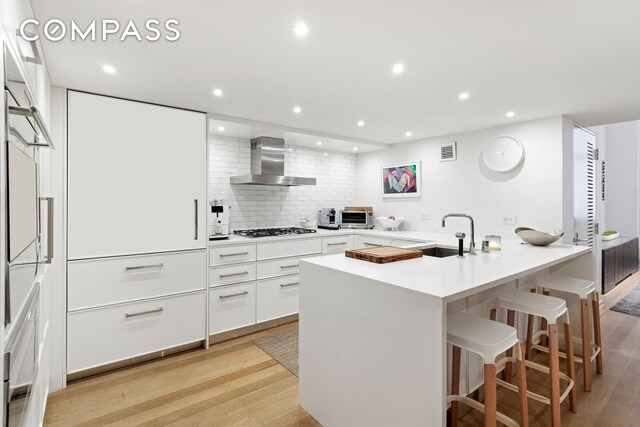 421 Hudson St Unit 221, New York City, NY 10014 - kitchen with wall chimney exhaust hood, gas stovetop, a sink, a kitchen bar, and white cabinets