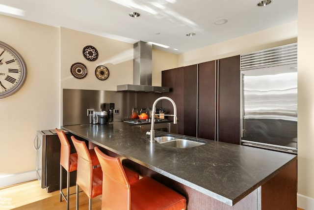 kitchen featuring a breakfast bar area, stainless steel appliances, dark brown cabinets, wall chimney exhaust hood, and sink