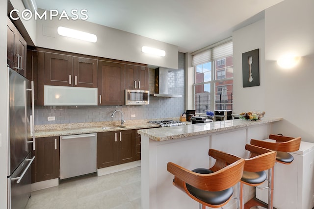 kitchen featuring dark brown cabinetry, stainless steel appliances, a sink, wall chimney range hood, and decorative backsplash