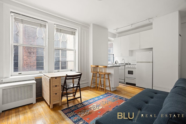 kitchen with a kitchen bar, radiator heating unit, white cabinetry, white appliances, and light wood-style floors