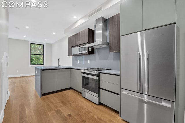 kitchen featuring wall chimney exhaust hood, built in appliances, sink, kitchen peninsula, and light wood-type flooring