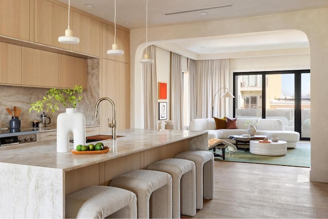 kitchen with light brown cabinets, hanging light fixtures, light wood-type flooring, light stone countertops, and decorative backsplash