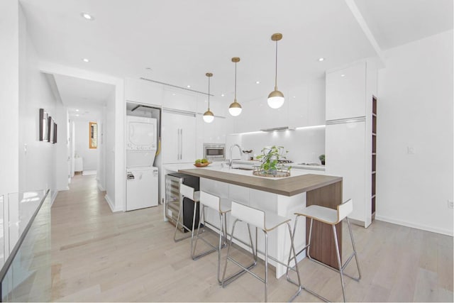 kitchen with light hardwood / wood-style flooring, stacked washer and clothes dryer, white cabinetry, hanging light fixtures, and a kitchen island with sink