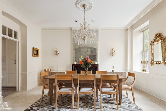 dining room with a wainscoted wall, light tile patterned flooring, crown molding, a chandelier, and a decorative wall