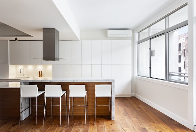 kitchen with modern cabinets, a wall mounted air conditioner, light countertops, wall chimney range hood, and a kitchen bar