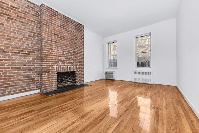 unfurnished living room featuring a fireplace, a wall unit AC, radiator, light wood-type flooring, and brick wall