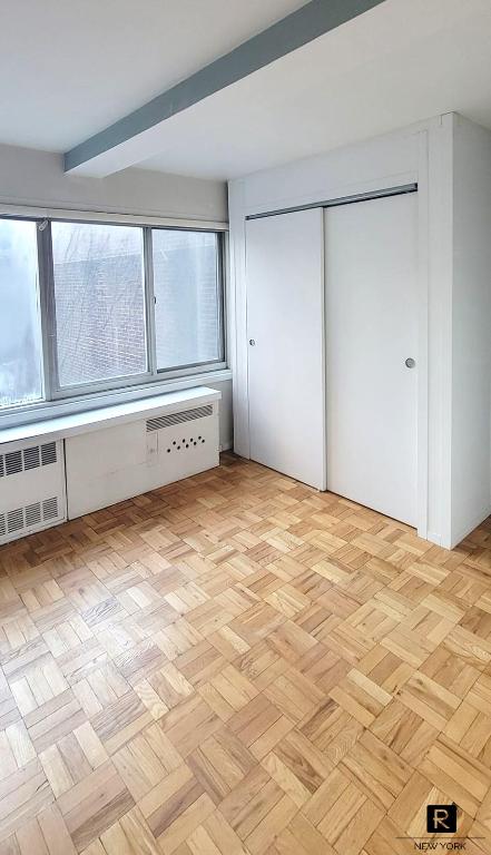 unfurnished bedroom featuring beamed ceiling, light parquet flooring, radiator heating unit, and a closet