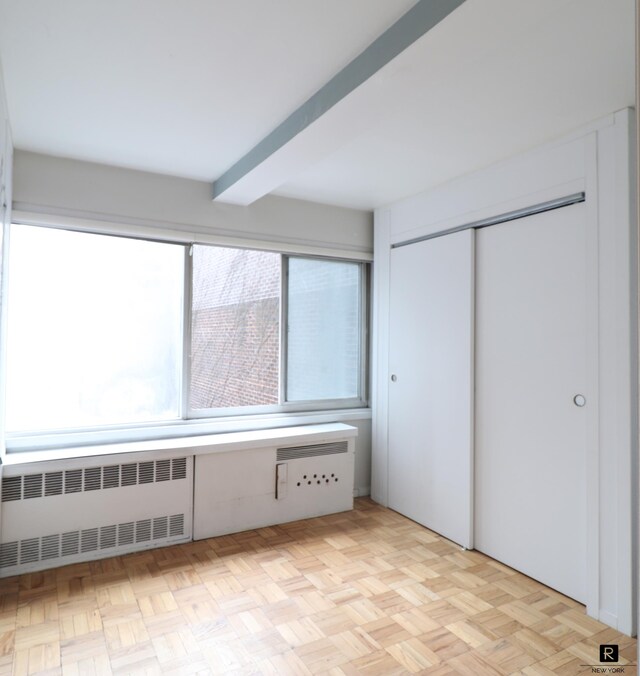 unfurnished bedroom featuring radiator, beam ceiling, a closet, and light parquet floors