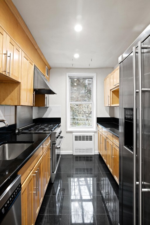 kitchen with high end appliances, dark countertops, granite finish floor, radiator heating unit, and under cabinet range hood