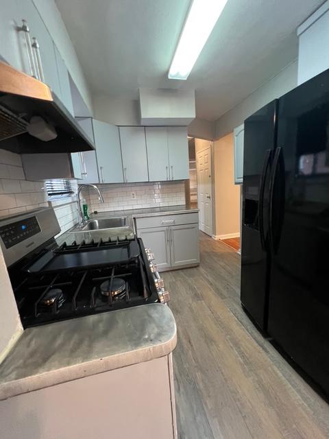 1241 E 98th St, New York City, NY 11236 - kitchen featuring black fridge with ice dispenser, light wood-type flooring, decorative backsplash, wall chimney exhaust hood, and a sink