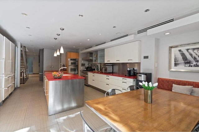 kitchen featuring wall chimney range hood, pendant lighting, sink, white cabinetry, and a kitchen island with sink