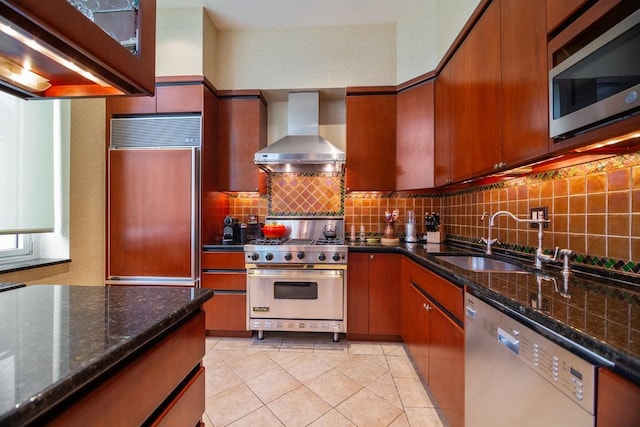 kitchen featuring backsplash, a sink, high quality appliances, dark stone counters, and wall chimney exhaust hood