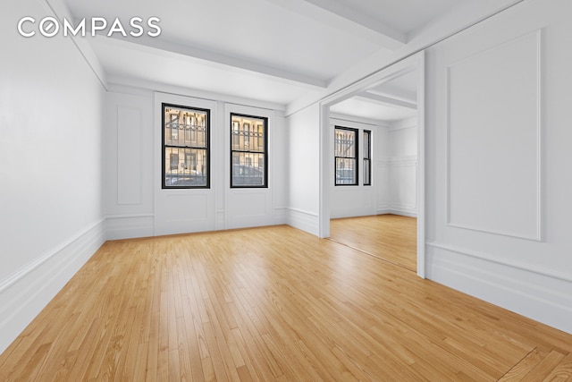 unfurnished room featuring baseboards, light wood finished floors, beamed ceiling, and a decorative wall