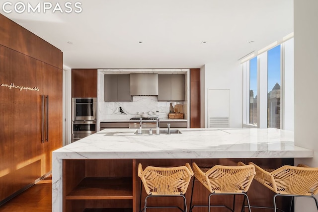 kitchen with backsplash, wall chimney range hood, floor to ceiling windows, a kitchen bar, and stainless steel double oven